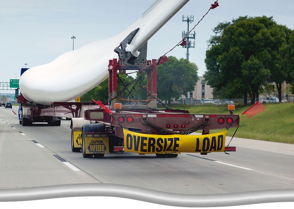 Large truck transporting a single wind turbine blade on the highway. Rear view, includes an "Oversize Load" banner on the back of the transport truck.