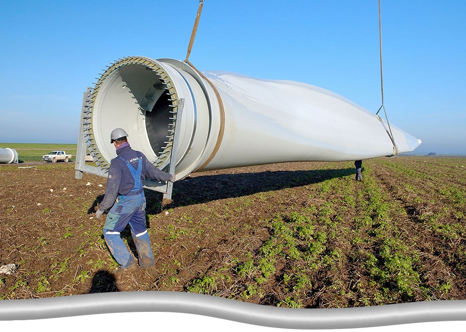 Worker in open farm field, guiding the movement/ positioning of a wind turbine blade, lifted by a crane (crane not shown in photo)